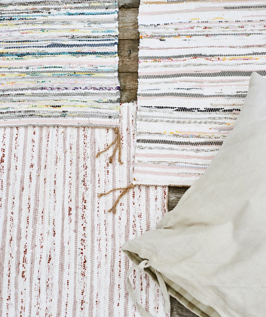 Three handwoven multicoloured striped rugs overlapping each other on wood decking, with a beige linen cushion on top.